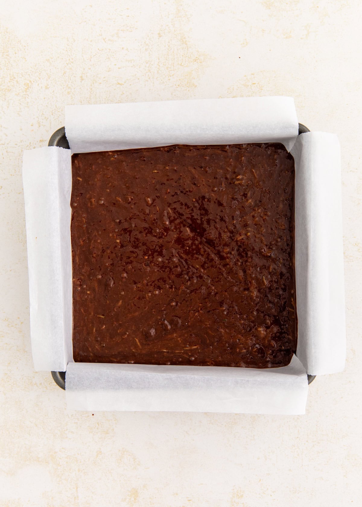 Brownie batter spread evenly in a parchment-lined square baking pan on a light surface.