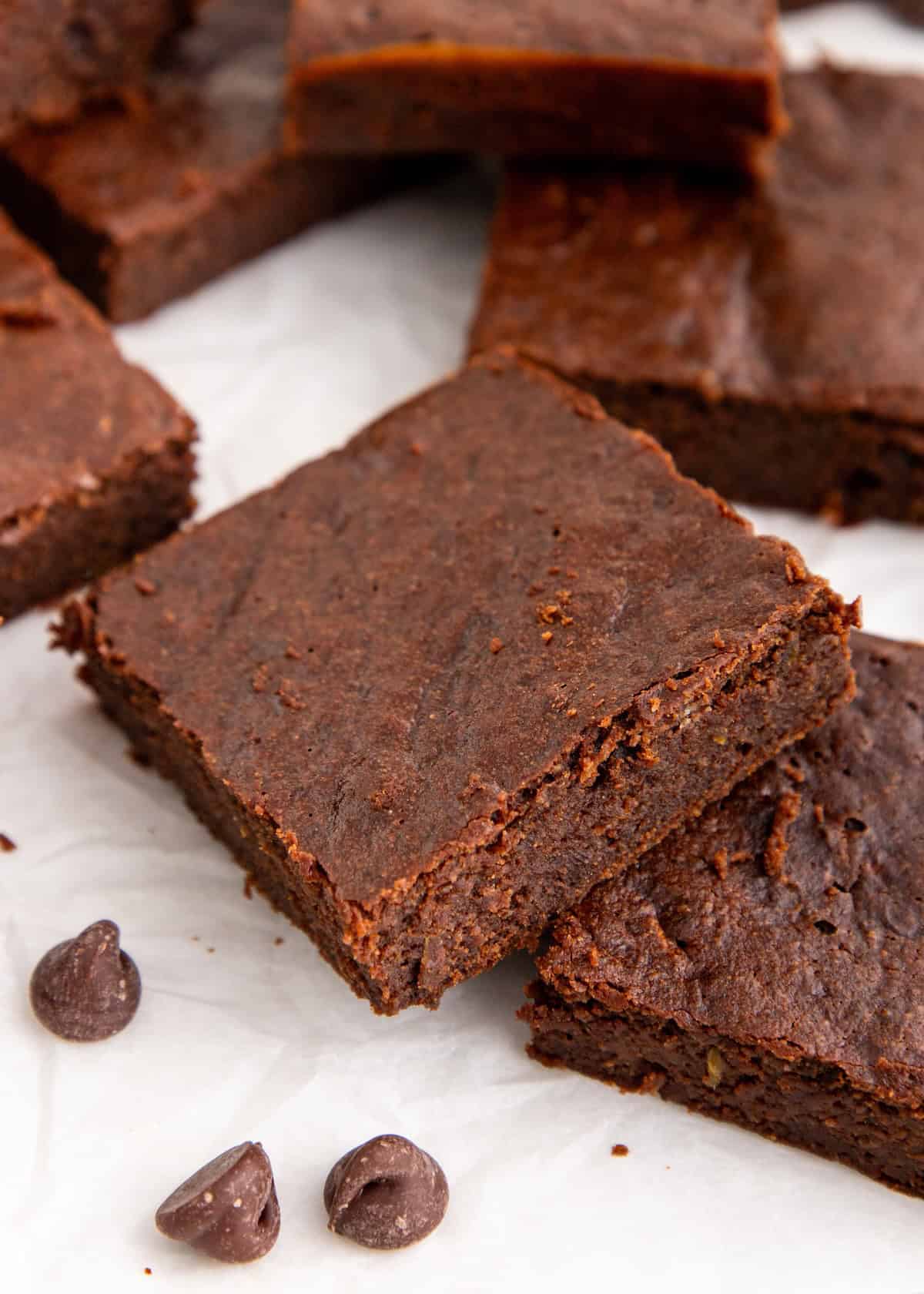 Fudgy brownies stacked on parchment paper with a few chocolate chips nearby.