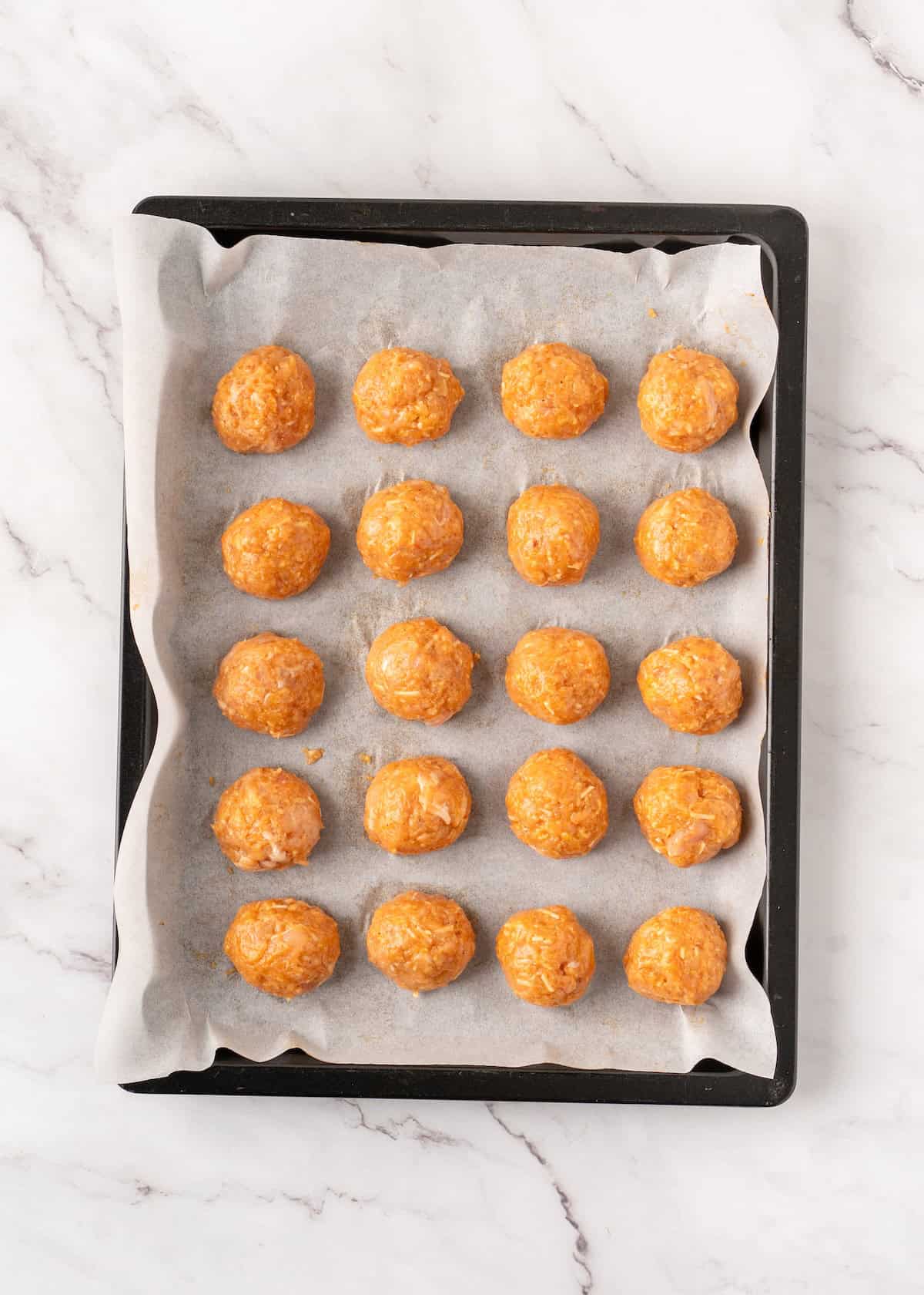 Twenty raw meatballs arranged in rows on a parchment-lined baking sheet.