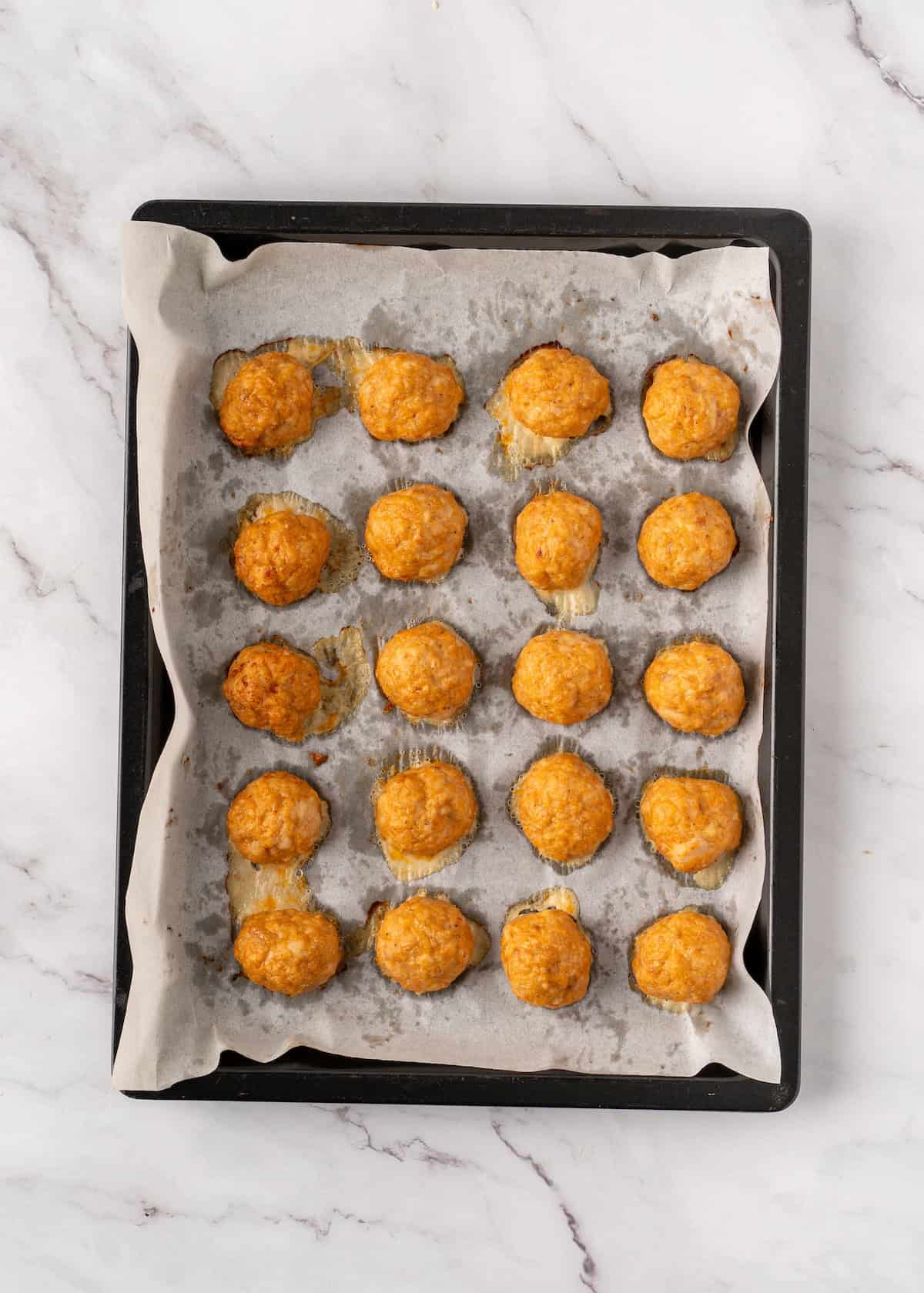 A baking sheet with twenty golden-brown meatballs on parchment paper.