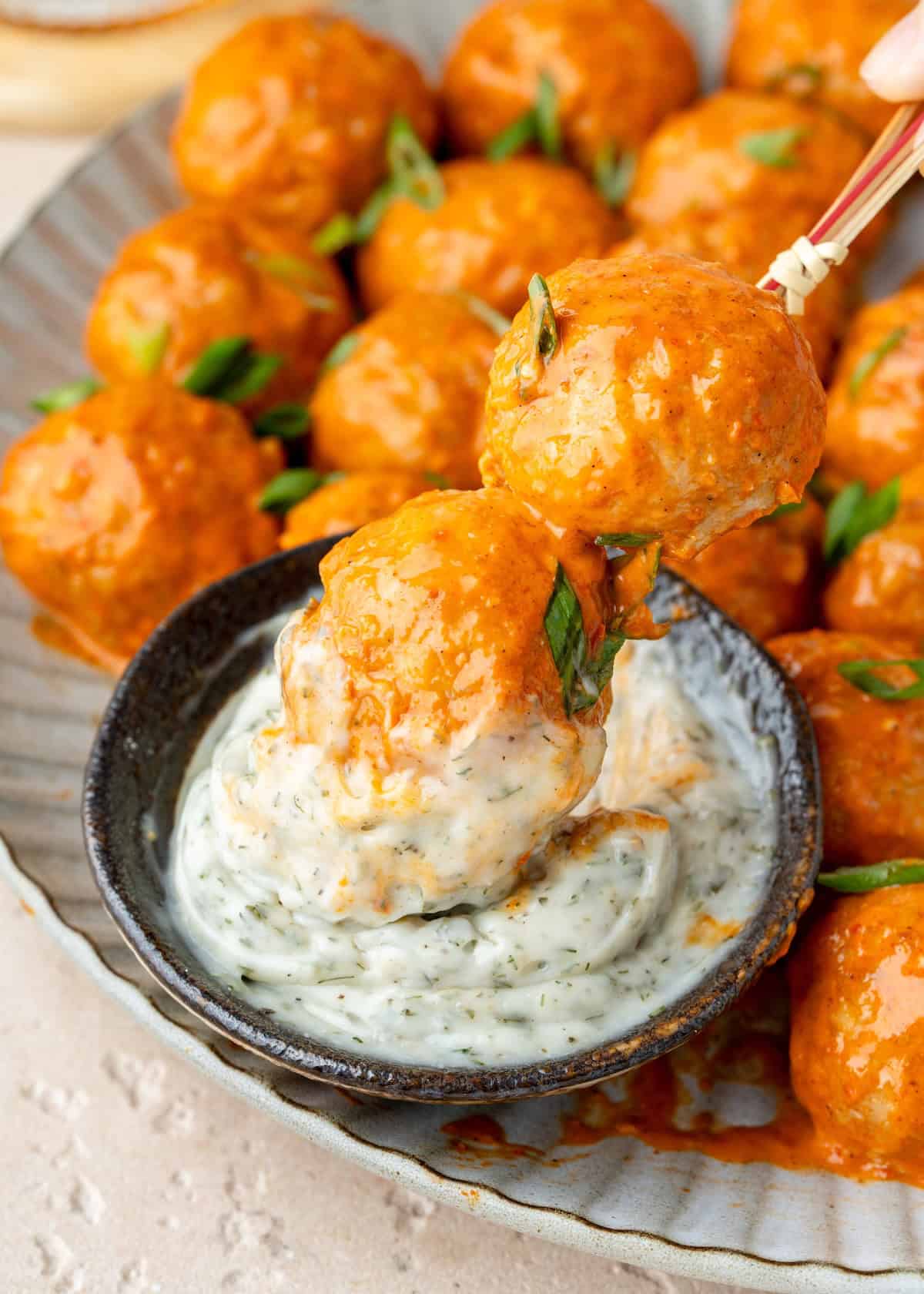 A skewer holding two buffalo meatballs dipped into a bowl of creamy dill sauce.